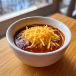 A white bowl filled with rich, dark red chili cheese dip, generously topped with shredded cheddar cheese.