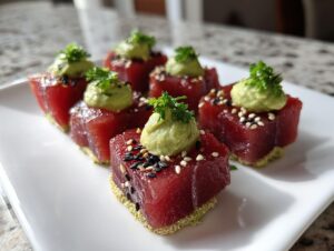 Six perfectly cubed pieces of vibrant red tuna tartare served on a white plate, topped with avocado puree and sesame seeds.