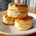 Three golden brown, fluffy buttermilk biscuits stacked on a white plate, glistening with a honey glaze.