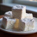 Close-up of several fluffy, square homemade marshmallows stacked on a white plate and heavily dusted with powdered sugar.