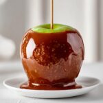 A glossy, thick caramel apple coated in rich, chewy caramel, sitting on a small white plate.