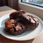 Three fudgy chocolate brownie cookies with cracked tops served on a white plate near a window.