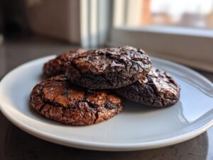 Close-up of several fudgy chocolate brownie cookies with cracked tops stacked on a light gray plate.