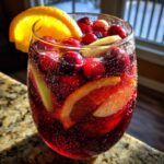 Close-up of a glass filled with bubbly, deep red Christmas sangria, loaded with fresh cranberries, orange slices, and apple chunks.