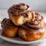 Close-up of stacked, gooey chocolate cinnamon rolls with thick chocolate frosting on a white plate.