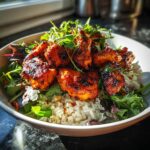 Close-up of a bowl featuring glistening, saucy harissa chicken pieces served over white rice and mixed greens.
