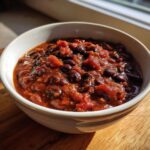 Close-up of a steaming bowl of rich, dark black bean chili with visible tomatoes, served on a wooden surface.