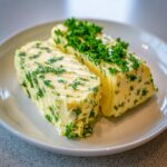 Two logs of homemade garlic butter mixed with vibrant green herbs, resting on a white plate.