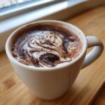 A close-up of rich homemade hot cocoa in a white mug, featuring a beautiful swirl of cream and chocolate syrup.