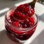 A spoonful of thick, vibrant red homemade raspberry jam being lifted from a glass jar.