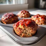 Four perfectly baked mini meatloaf portions topped with a shiny glaze in a metal muffin tin.