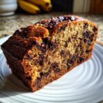 A thick, moist slice of chocolate chip banana bread sits on a white plate, showing rich chocolate chunks throughout.