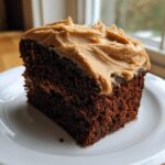 A close-up of a moist slice of dark gingerbread cake topped with thick, swirled brown frosting on a white plate.
