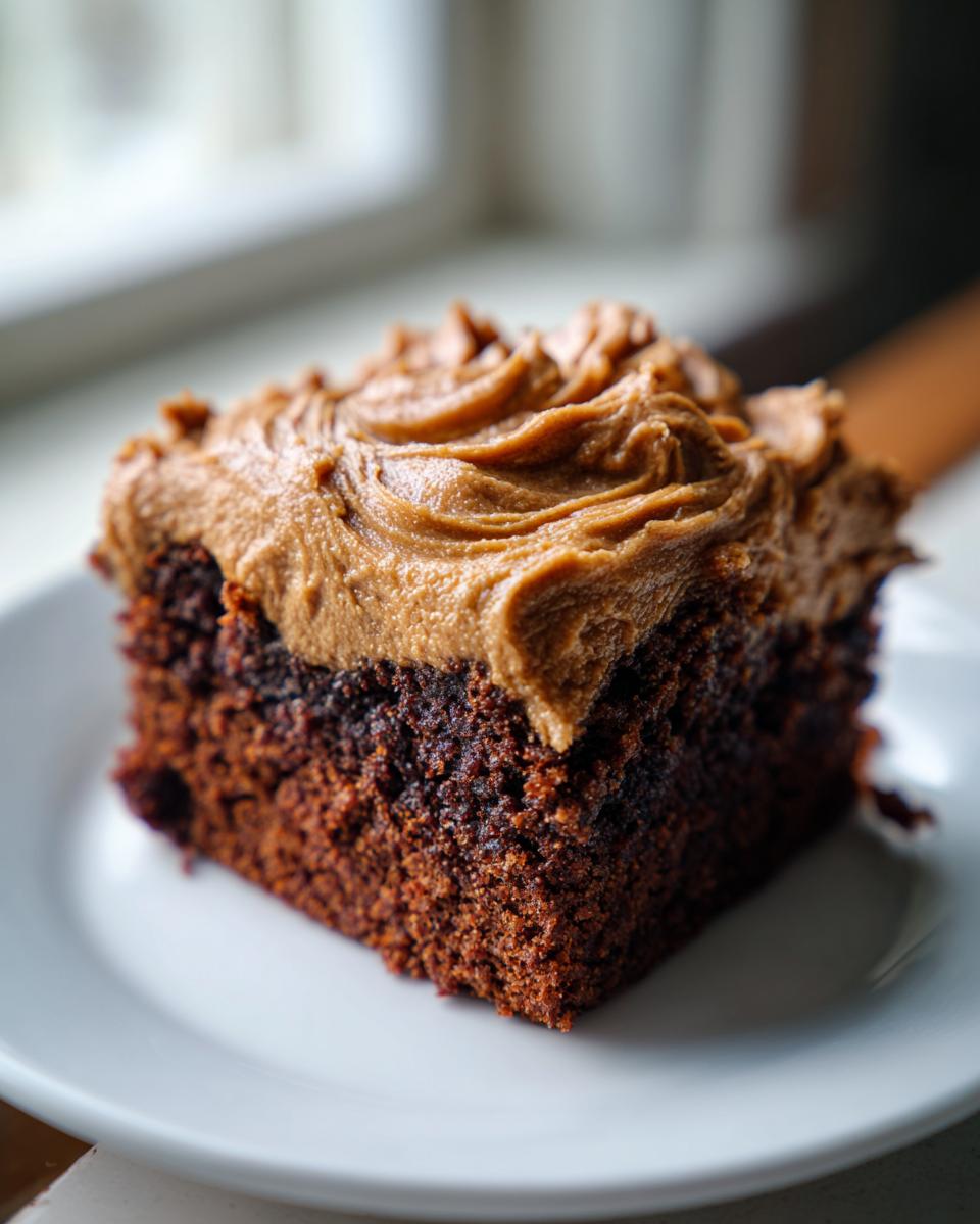 Close-up of a square slice of moist, dark gingerbread cake topped with thick, swirled brown frosting on a white plate.