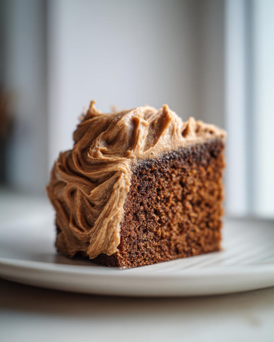 Close-up of a moist slice of gingerbread cake topped with thick, swirled brown frosting on a white plate.