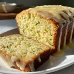 Close-up of a loaf of lemon zucchini bread, sliced, showing shredded zucchini and a thick lemon glaze dripping down the sides.