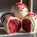 Close-up of moist red velvet cake pops, one cut in half showing the rich red interior and white chocolate coating.