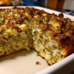 Close-up of a freshly baked, moist sausage stuffing with a golden-brown, herbed top, showing a serving removed.