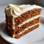 Close-up of a moist, three-layer spice cake slice topped with thick cream cheese frosting swirls.