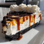 A decadent slice of no-bake ice cream pie featuring a dark cookie crust, creamy filling, caramel drizzle, and whipped cream topping.