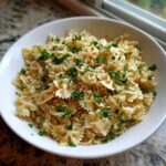 A white bowl filled with a mix of rice pilaf and bowtie pasta, topped generously with fresh chopped parsley.