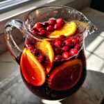 A glass pitcher filled with deep red holiday sangria, floating cranberries, and bright orange slices.