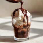 A spoonful of rich chocolate syrup is being poured over scoops of vanilla ice cream in a glass.
