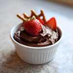 A serving of rich, creamy chocolate dip in a white ramekin, topped with a whole strawberry and served with pretzels.