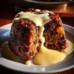 A close-up of a rich, dark slice of plum pudding covered in creamy vanilla custard sauce.