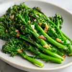 Close-up of bright green roasted broccolini stalks topped with caramelized garlic pieces on a light plate.