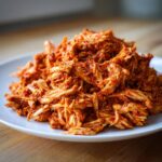 A close-up of shredded chicken tinga piled high on a white plate, coated in rich red sauce.