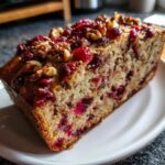 A thick slice of moist cranberry walnut bread, topped with whole walnuts and bright red cranberries, sitting on a white plate.