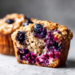 A close-up view of a soft blueberry oatmeal muffin cut in half, showing moist crumb and burst blueberries.