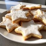 Close-up of star-shaped vegan sugar cookies piled on a white plate, topped with shiny white icing and sprinkles.
