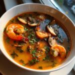 A close-up of a white bowl filled with vibrant, orange-red tom yum soup featuring large shrimp and sliced mushrooms, garnished with fresh herbs.