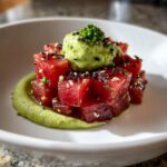 Close-up of stacked, ruby-red tuna tartare cubes topped with avocado puree and black sesame seeds.