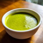 Close-up of a creamy, vibrant green zucchini soup served in a small white bowl on a wooden table.