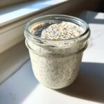A glass jar filled with creamy, prepared chia pudding sitting on a white surface near a window.