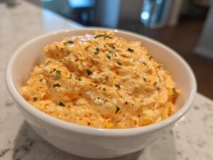 Close-up of creamy, orange-colored boat dip topped with green herbs in a white serving bowl.