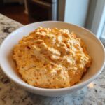 A close-up of creamy, orange-colored boat dip piled high in a white bowl on a granite counter.