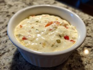 Close-up of a creamy, warm cheese dip with visible red and green peppers served in a small white ramekin.