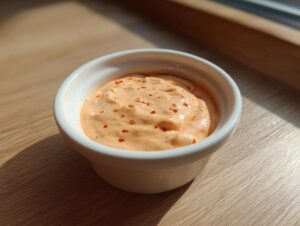 A close-up of creamy, orange-hued smash burger sauce sprinkled with red pepper flakes in a small white bowl.