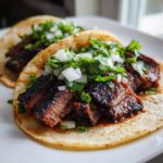 Close-up of two delicious steak tacos on corn tortillas topped with charred steak, diced white onion, and fresh cilantro.