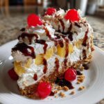 A tall slice of banana split cake featuring layers of whipped cream, pineapple chunks, and a graham cracker crust, topped with chocolate syrup and maraschino cherries.