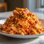 A generous mound of vibrant orange buffalo chicken salad mixed with green celery and red onion pieces on a white plate.