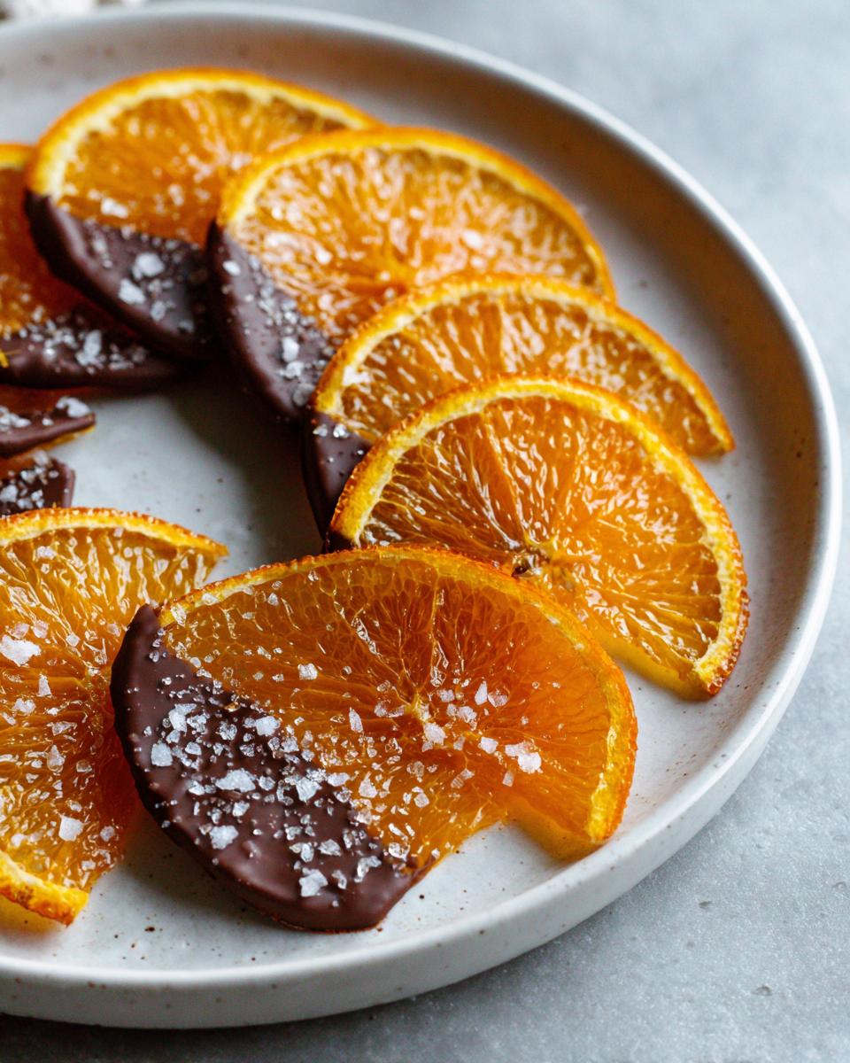 Close-up of glistening candied orange slices, partially dipped in dark chocolate and sprinkled with flaky sea salt.