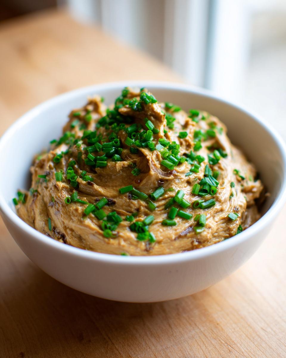 A close-up of creamy caramelized onion dip in a white bowl, garnished heavily with fresh chopped chives.