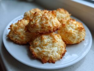 A close-up of several golden brown, chewy coconut cookies stacked on a white plate.