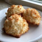 Three golden brown, chewy coconut cookies piled on a white plate near a window.