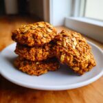 A stack of four chewy pumpkin oatmeal cookies topped with visible oats, resting on a white plate.